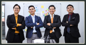 Four male professionals in blazers standing together with crossed arms and confident smiles in front of a whiteboard with charts.