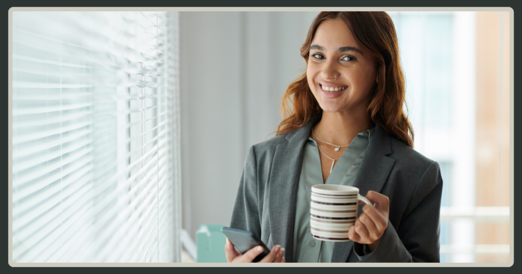 A smiling young woman in a gray business jacket holding a striped coffee mug and a phone while standing by a window with blinds.