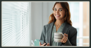A smiling young woman in a gray business jacket holding a striped coffee mug and a phone while standing by a window with blinds.