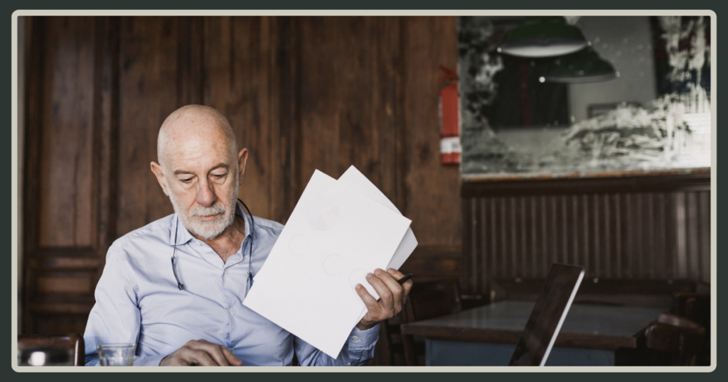 An older man with a white beard sitting at a wooden table, carefully reviewing a stack of white papers held in his hand.