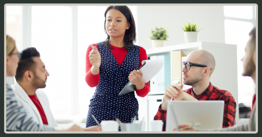 A woman in a polka-dot dress standing and leading a group discussion with several seated colleagues in a bright office, holding a clipboard.