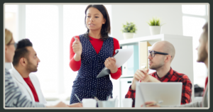 A woman in a polka-dot dress standing and leading a group discussion with several seated colleagues in a bright office, holding a clipboard.