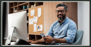 A smiling man with a grey-streaked beard and glasses sits at an office desk holding a smartphone, with a computer and data-filled corkboard behind him.