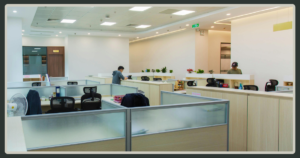 A wide-angle view of a bright, modern open-plan office featuring several rows of clean, light-colored cubicles and workstations.