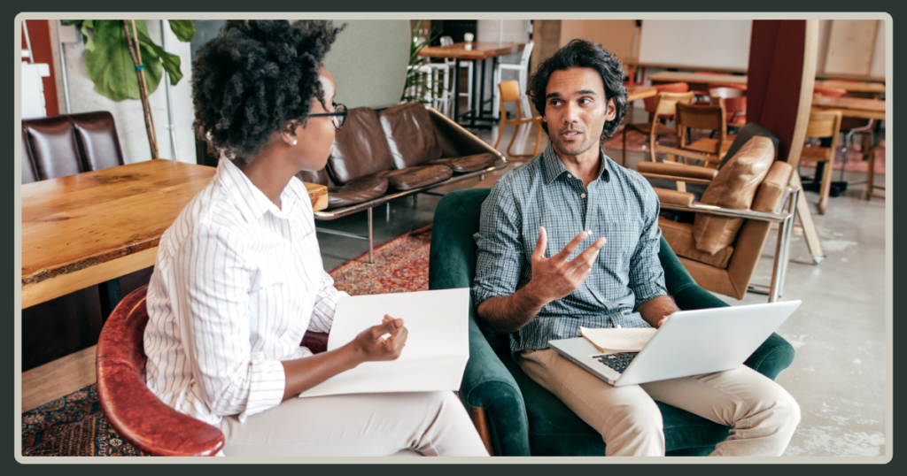 A man gesturing expressively while speaking to a colleague in a relaxed office lounge, representing a mentorship or coaching conversation.