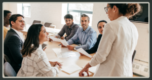 A group of professionals sitting at a conference table, smiling and listening intently to a female leader presenting at the head of the table.