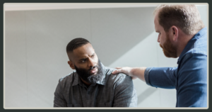 A supportive moment in the office where one man places a comforting, empathetic hand on his colleague's shoulder during a serious conversation.