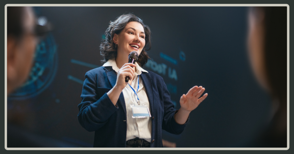 A woman in a professional blazer speaking confidently into a microphone with a bright smile during a leadership training event.