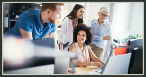A diverse group of four colleagues smiling while gathered around a computer monitor to collaborate on a digital project.