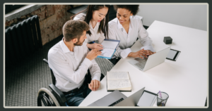 A collaborative team, including a man in a wheelchair and two women, reviewing notes and a laptop together at a white office desk.