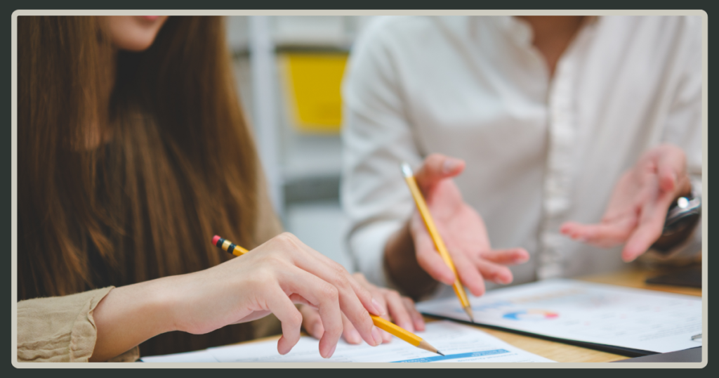 A close-up shot of two people's hands using pencils to review printed data charts and performance reports on a wooden desk.