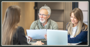 An older man with grey hair and glasses holds a document while leading an earnest discussion with two female colleagues at a conference table.