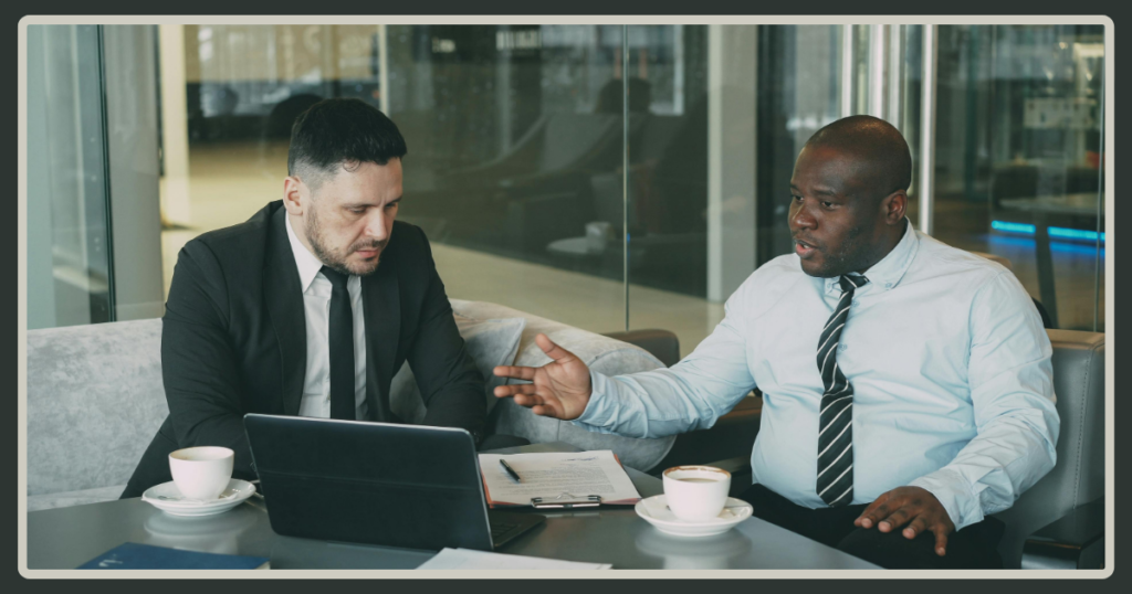 A Black man in a light blue shirt gestures expressively while discussing a document with a colleague in a black suit sitting on a grey couch.