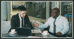 A Black man in a light blue shirt gestures expressively while discussing a document with a colleague in a black suit sitting on a grey couch.