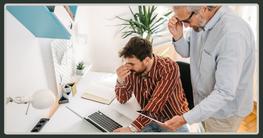 A man in a striped shirt looks stressed at his desk while an older manager stands over his shoulder, peering closely at his tablet.
