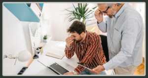 A man in a striped shirt looks stressed at his desk while an older manager stands over his shoulder, peering closely at his tablet.