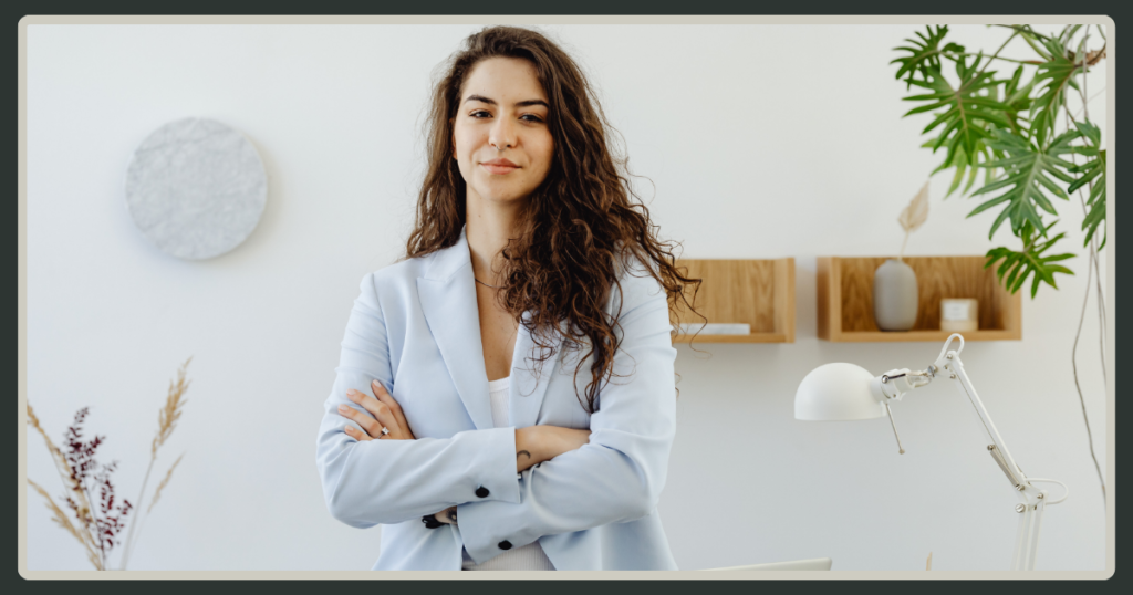A woman with long curly hair wearing a light blue blazer stands with her arms crossed, smiling confidently in a clean, minimalist office setting.