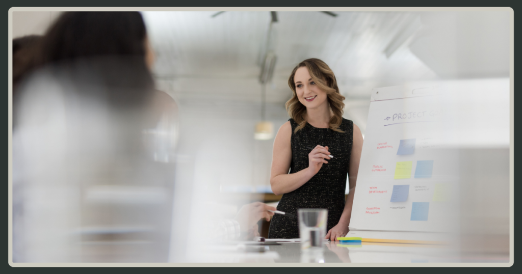 A woman in a black dress stands next to a whiteboard with colorful sticky notes, smiling as she leads a strategic planning session.