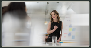 A woman in a black dress stands next to a whiteboard with colorful sticky notes, smiling as she leads a strategic planning session.