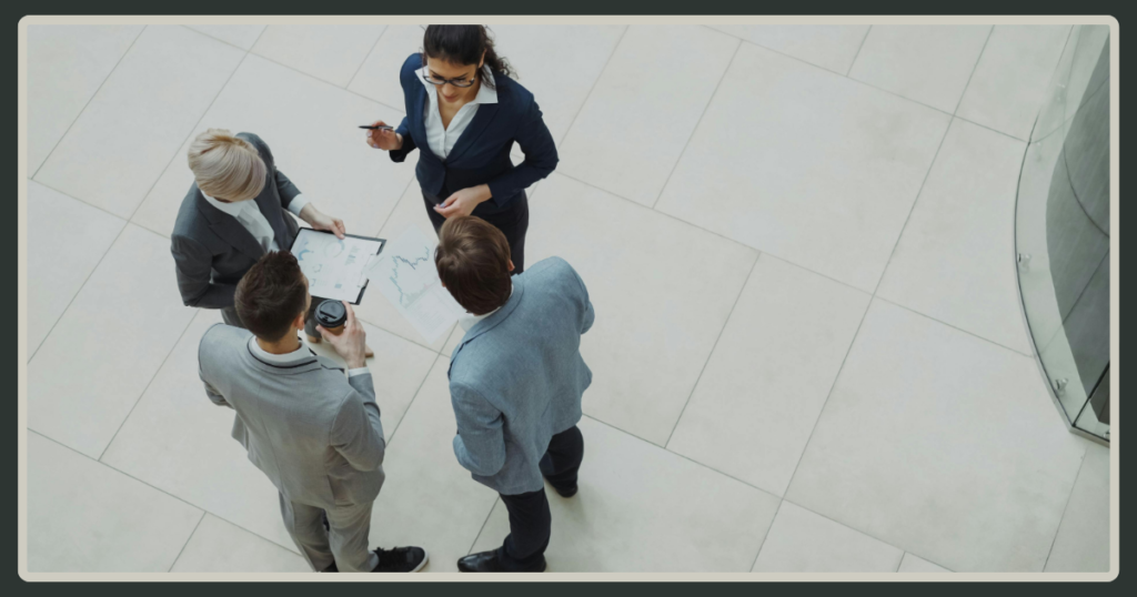 A top-down, high-angle view of four business professionals in suits standing in a circle in a bright office atrium, reviewing printed charts together.