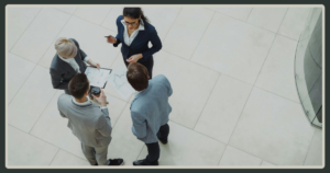 A top-down, high-angle view of four business professionals in suits standing in a circle in a bright office atrium, reviewing printed charts together.