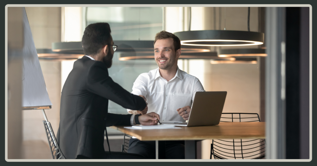 Two men in professional attire shaking hands across a wooden office desk with an open laptop, symbolizing a successful partnership or new hire.