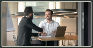 Two men in professional attire shaking hands across a wooden office desk with an open laptop, symbolizing a successful partnership or new hire.