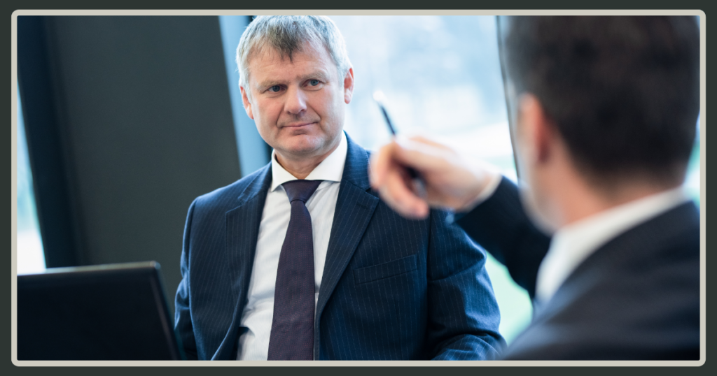 A close-up shot of a senior businessman in a dark pinstripe suit listening intently to a colleague who is gesturing toward him with a pen.