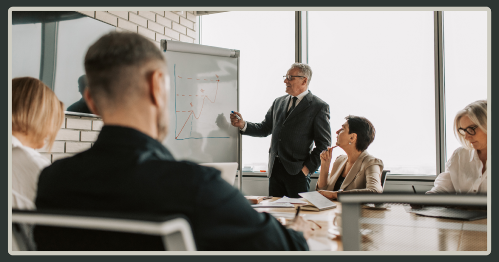 A senior professional standing at a whiteboard, pointing to a growth line graph while presenting to a seated team in a sunlit conference room.