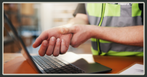 A close-up of a worker wearing a high-visibility safety vest, holding their wrist in pain while sitting at a desk with an open laptop.