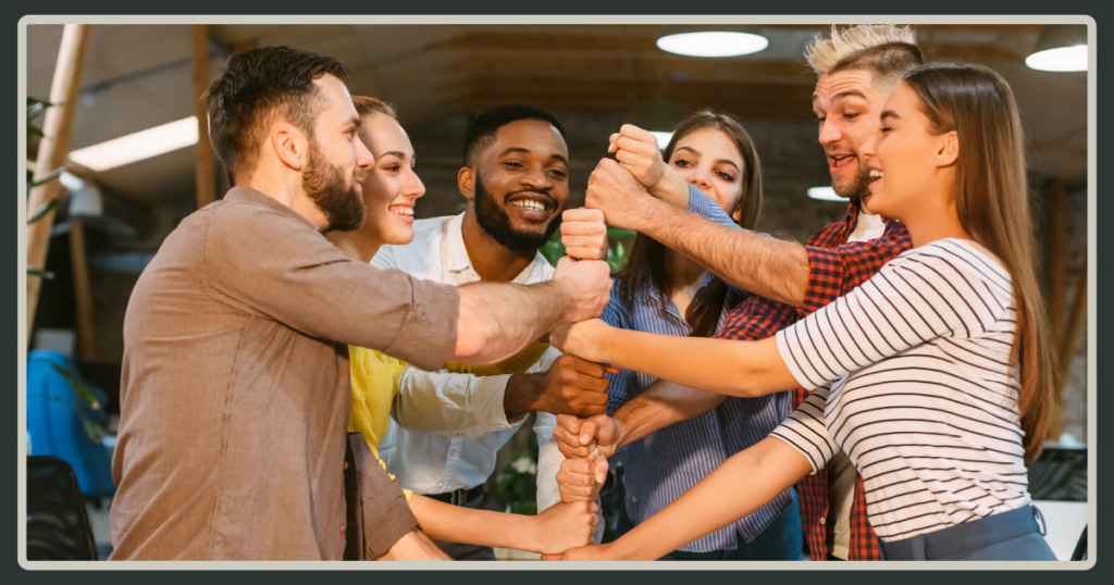 A diverse group of smiling coworkers stacking their fists together in a central column, celebrating a team achievement in a modern office.