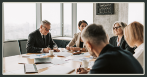 A group of senior business leaders sitting around a large wooden table with focused, serious expressions while reviewing documents.