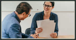 A smiling woman with glasses and a polka-dot shirt holding a stack of resumes while talking to a man during a job interview.