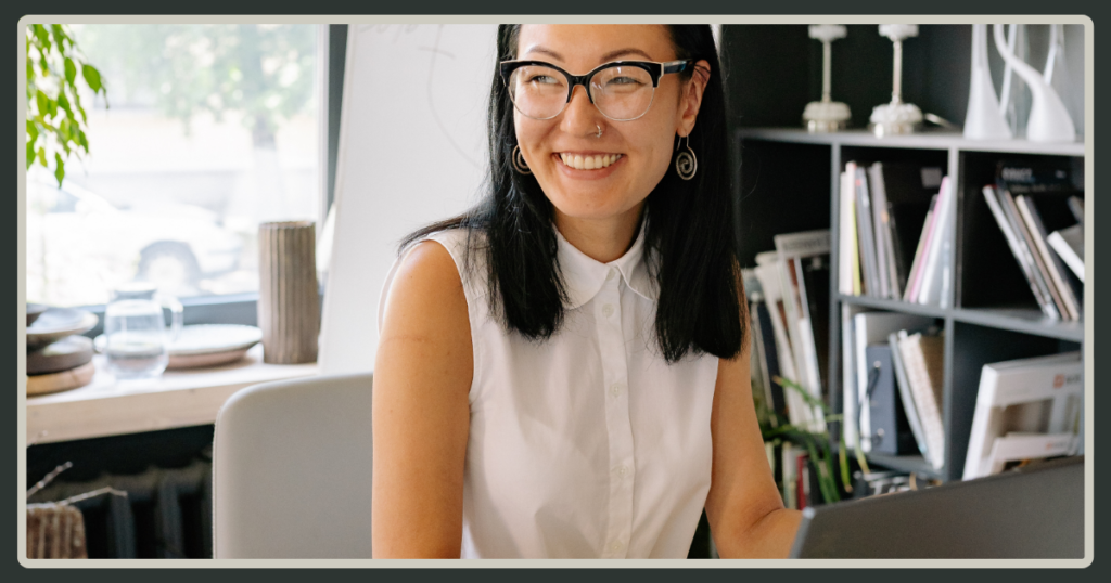 A smiling woman with glasses and dark hair sitting at an office desk with a laptop, looking cheerfully to the side in a bright workspace.